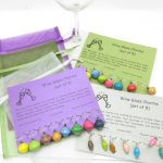 Photo with 3 sets of wine glass charms on a different color of informative card. Each card contains a set of 8 wine charm beads in different colors. 3 organza gift bags are shown behind the cards.