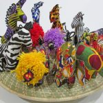 Assorted small miniature stuffed animals in 5 animal shapes of elephant, giraffe, lion, zebra and rhino. the animals are displayed on a woven grass looking circular shape. The animals come in a variety of bright colors except the zebra is black and white.