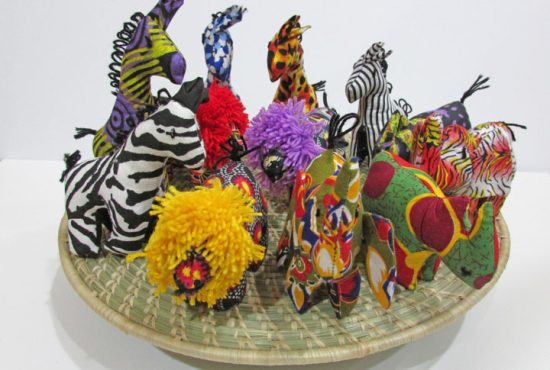 Assorted small miniature stuffed animals in 5 animal shapes of elephant, giraffe, lion, zebra and rhino. the animals are displayed on a woven grass looking circular shape. The animals come in a variety of bright colors except the zebra is black and white.