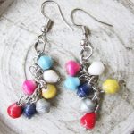 One pair of multi-color mini bead dangle earrings on small hook laying on a quartz-like surface in swirling gray on white. Each earring contains about 7 tiny colorful beads. Handmade by Ugandan women artisans from recycled paper.