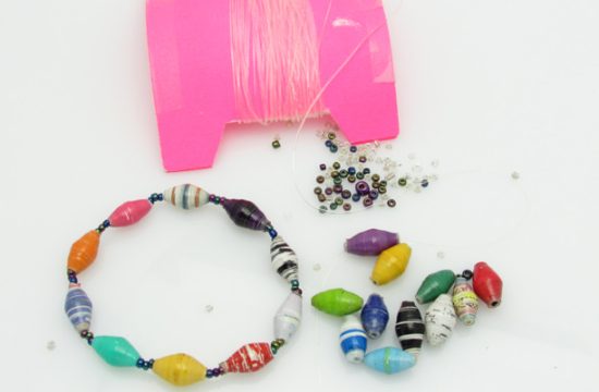 Bead bracelet making kit shown with all its parts including stretch bead cord, tiny seed beads and paper beads, plus an example of the finished product: a multicolor beaded bracelet. This kit makes one bracelet and is available for purchase.