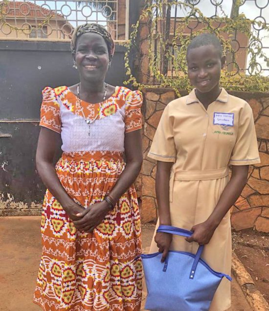 Secondary student in Jinja, in school uniform, standing with her smiling mother, and holding a gift from her sponsor in the U.S.